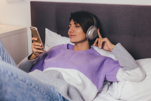 Donna felice a casa sul letto comodo indossando pullover vestiti caldi, ascoltare musica