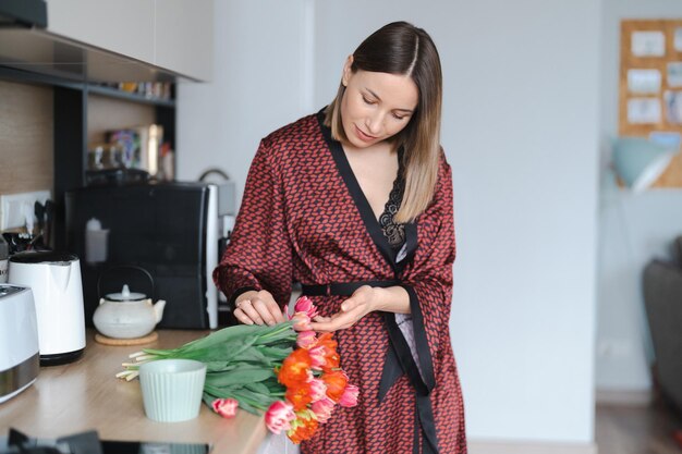 Donna felice che beve un caffè a casa in cucina indossando un abito di seta mentre si gustano fiori freschi