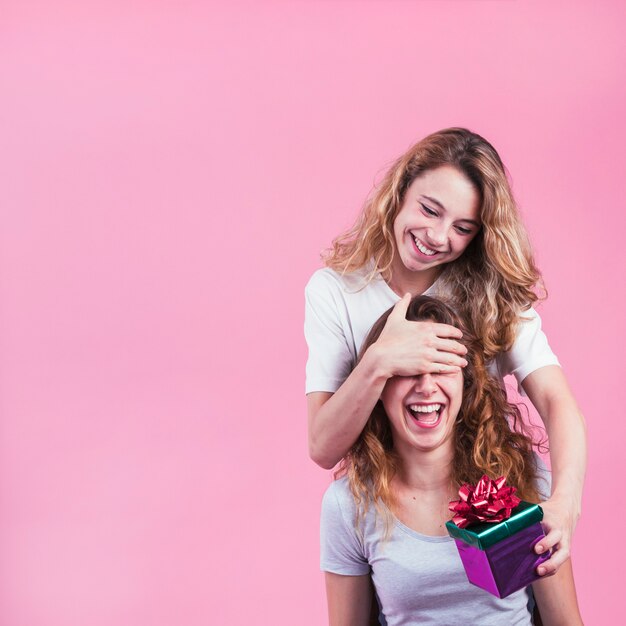 Donna felice che copre i suoi occhi femminili che tengono il contenitore di regalo contro fondo rosa