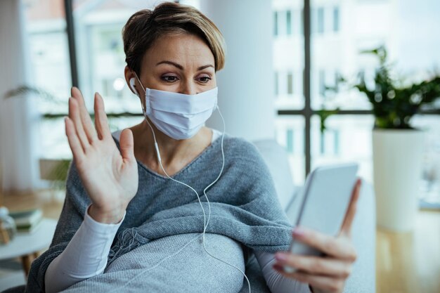 Donna felice che indossa la maschera facciale N95 e ha una chat video sul telefono cellulare e saluta qualcuno