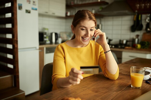 Donna felice che parla al telefono mentre si utilizza la carta di credito a casa