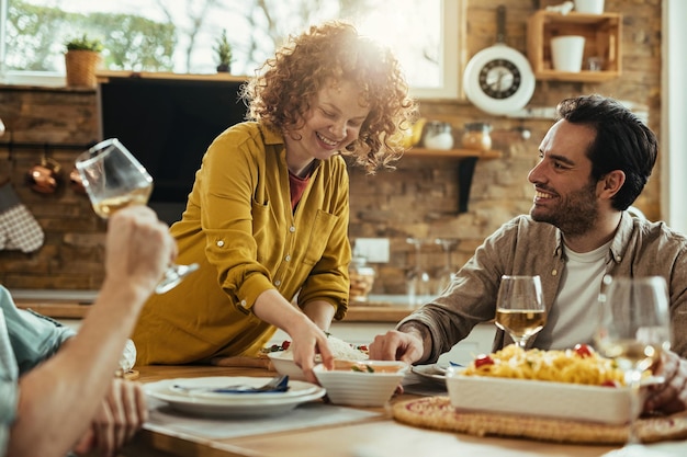 Donna felice che porta cibo a tavola e si diverte con i suoi amici durante l'ora di pranzo a casa