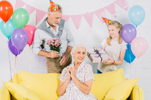 Donna felice che si siede sul sofà davanti al marito e alla nipote che tengono i regali di compleanno