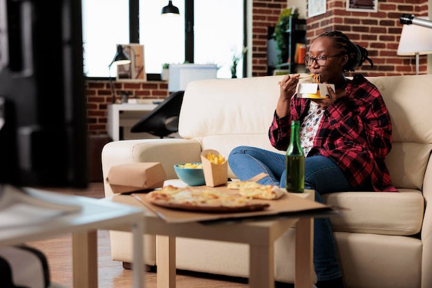 Donna felice che usa le bacchette per mangiare le tagliatelle dalla scatola di consegna, godendosi il pasto davanti alla televisione che guarda film. Mangiare un pacchetto da asporto di fast food come cena, guardare un film in tv.