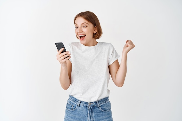 Donna felice che vince sul videogioco per smartphone alzando la mano e tifando gridando sì con gioia raggiungere l'obiettivo online in piedi su sfondo bianco