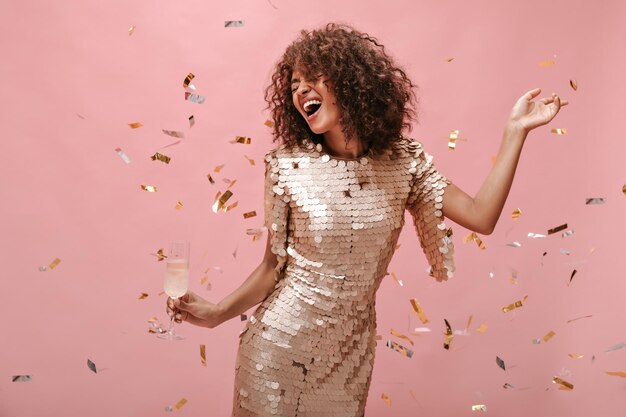 Donna felice con capelli scuri ondulati in abiti alla moda lucidi che si rallegrano tenendo il bicchiere con champagne e posando con coriandoli su sfondo rosa