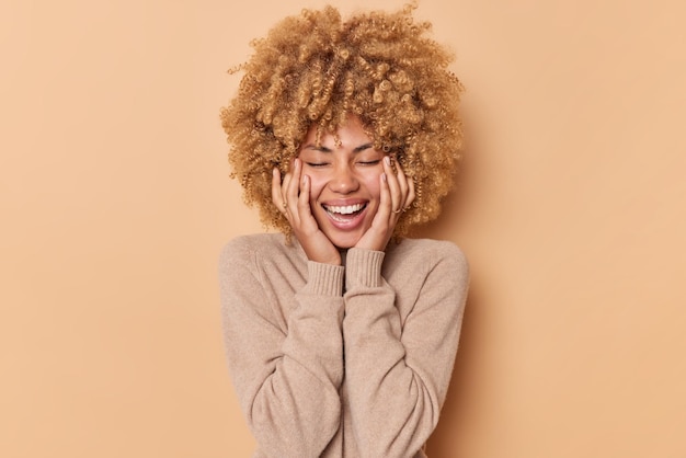 Donna felice con i capelli ricci tiene le mani sulle guance chiude gli occhi e sorride con gioia indossa un maglione casual essere di buon umore isolato su sfondo beige richiama bei ricordi. Concetto di emozioni