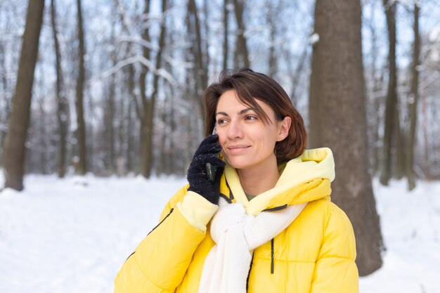 Donna felice di ottimo umore cammina attraverso la foresta invernale innevata e chiacchiera allegramente al telefono, godendosi il tempo all'aperto nel parco