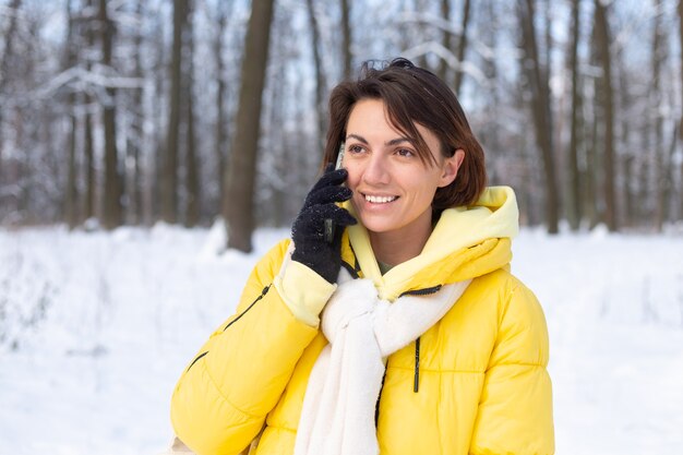 Donna felice di ottimo umore cammina attraverso la foresta invernale innevata e chiacchiera allegramente al telefono, godendosi il tempo all'aperto nel parco