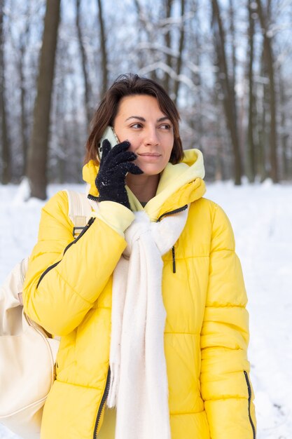 Donna felice di ottimo umore cammina attraverso la foresta invernale innevata e chiacchiera allegramente al telefono, godendosi il tempo all'aperto nel parco