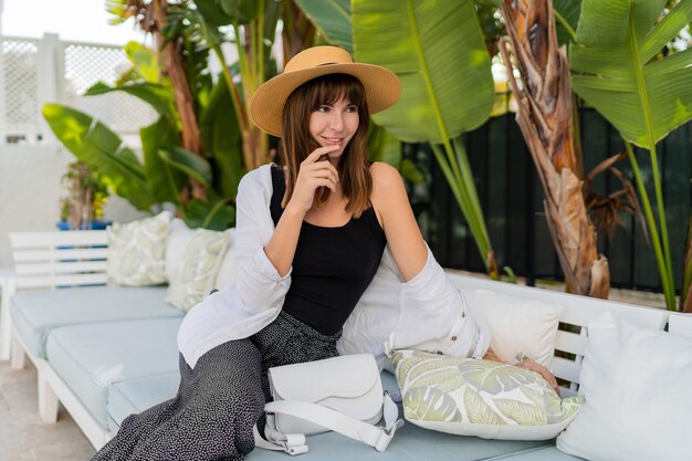 Donna felice in cappello di paglia agghiacciante a casa, sulla terrazza di lusso, in posa vicino al giardino tropicale.