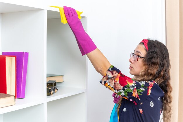 Donna felice in guanti di gomma che pulisce gli scaffali bianchi con uno straccio giallo usando uno spray detergente in casa in un soggiorno luminoso