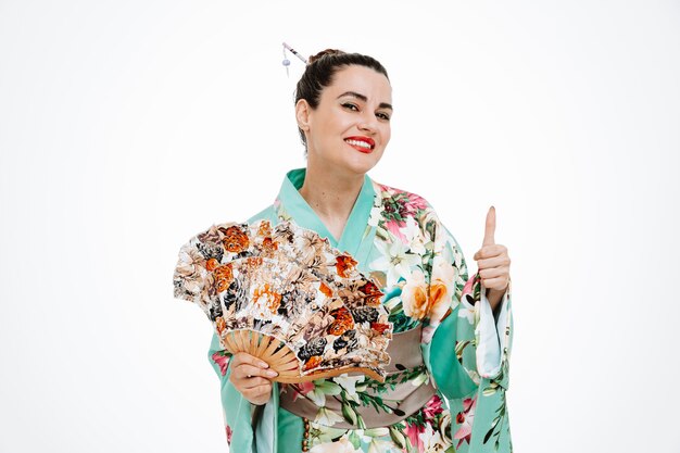 Donna felice in kimono giapponese tradizionale che tiene ventaglio sorridente che mostra ampiamente il segno ok su bianco