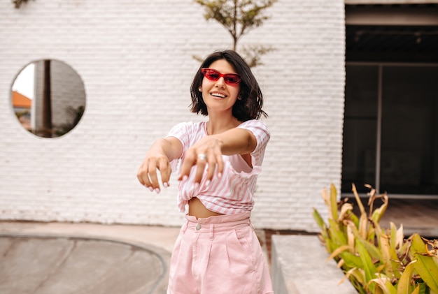 Donna felice in occhiali da sole in posa sulla strada con le mani tese. Colpo esterno di donna abbronzata in maglietta rosa.