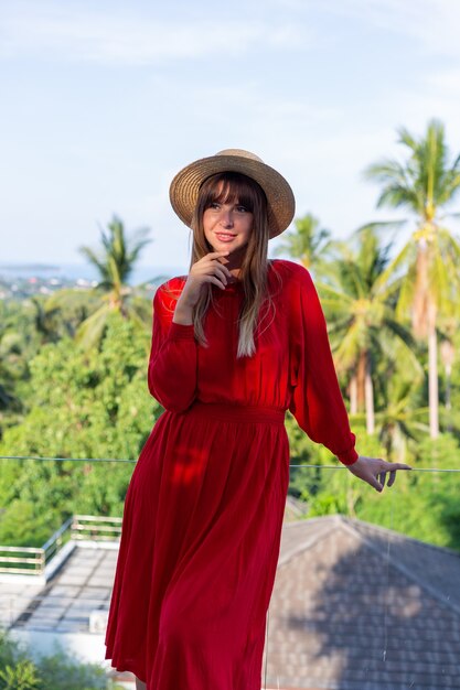 Donna felice in vacanza in abito estivo rosso e cappello di paglia sul balcone con vista tropicale sul mare e alberi di plam.