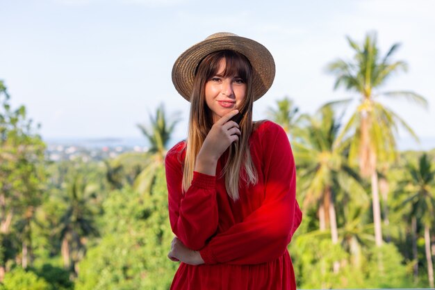 Donna felice in vacanza in abito estivo rosso e cappello di paglia sul balcone con vista tropicale sul mare e alberi di plam.
