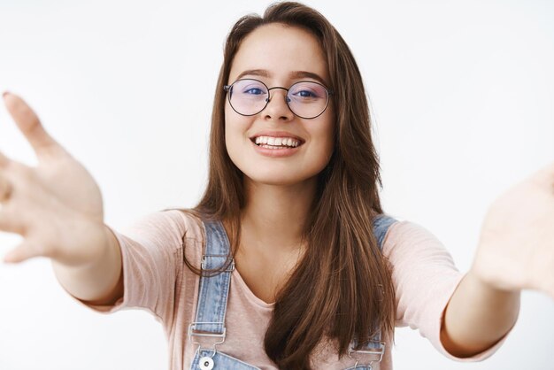 Donna femminile tenera e attraente premurosa con gli occhiali che estendono le braccia verso la telecamera per dare baci e abbracci volendo tenersi tra le braccia cosa sorridente ampiamente in piedi felice e carina sul muro grigio