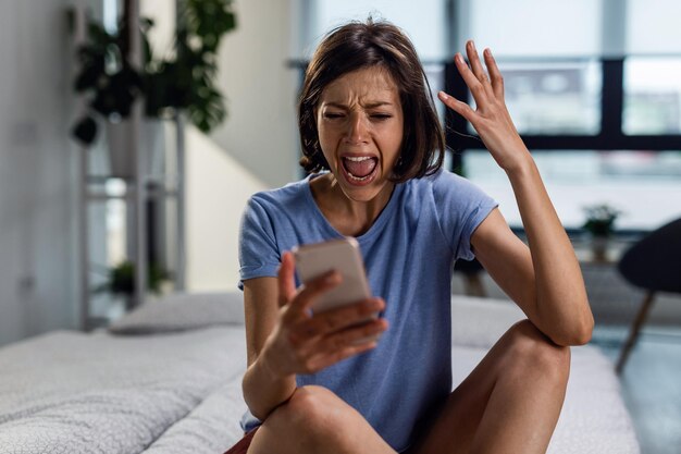 Donna frustrata che grida al suo telefono mentre è seduta sul letto e legge il messaggio di testo che ha ricevuto