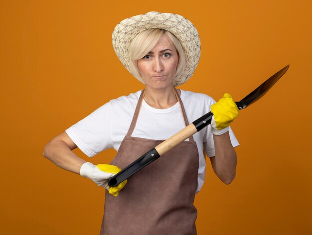 Donna giardiniere di mezza età accigliata in uniforme da giardiniere che indossa cappello e guanti da giardinaggio che tengono vanga guardando la parte anteriore isolata sul muro arancione