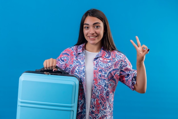 Donna giovane bella viaggiatore che tiene la valigia blu sorridente allegramente facendo segno di vittoria pronto a viaggiare in piedi su sfondo blu