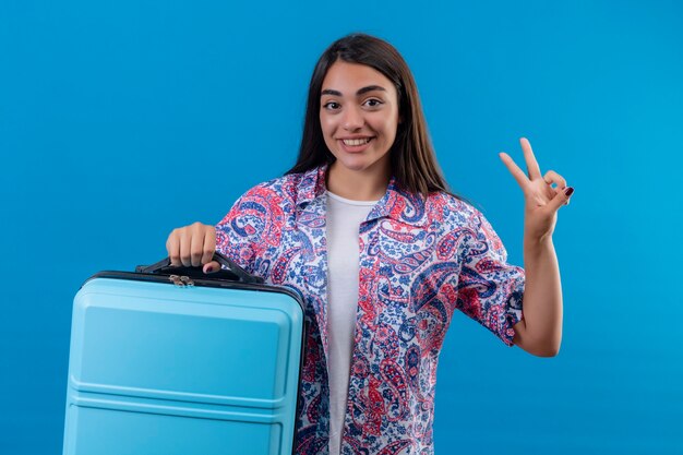 Donna giovane bella viaggiatore che tiene la valigia blu sorridente allegramente facendo segno di vittoria pronto a viaggiare in piedi su sfondo blu