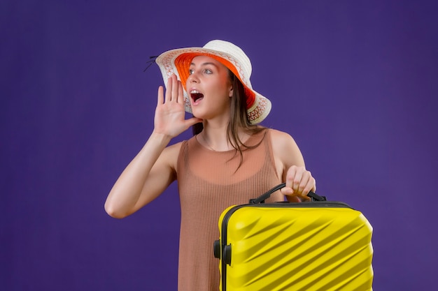 Donna giovane bella viaggiatore in cappello estivo con valigia gialla che grida o chiama qualcuno con la mano vicino a falena in piedi su sfondo viola