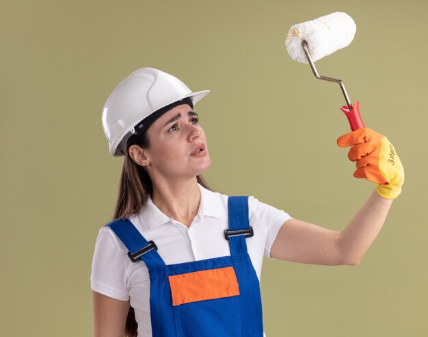 Donna giovane costruttore di pensiero in uniforme e guanti che alzano e che esaminano la spazzola del rullo isolata sulla parete verde oliva