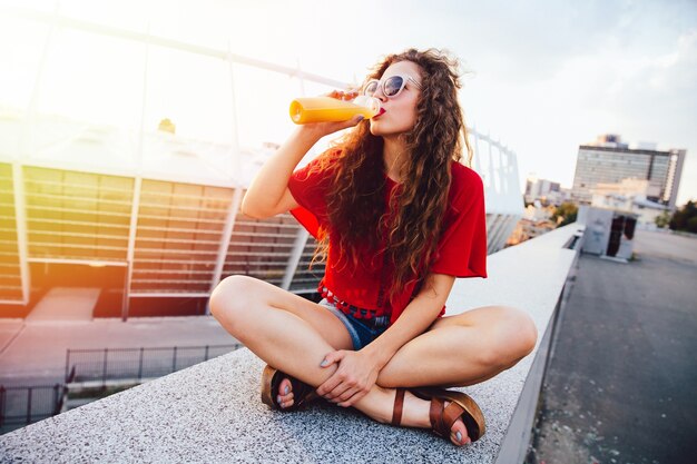 Donna giovane hipster con i capelli ricci in occhiali da sole bere succo d&#39;arancia fresco dalla bottiglia
