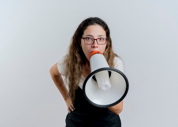 Donna giovane parrucchiere professionista in grembiule che grida al megafono ad alta voce in piedi sopra il muro bianco