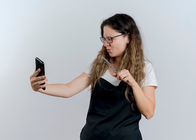 Donna giovane parrucchiere professionista in grembiule che tiene le forbici guardando il suo schermo dello smartphone essendo scontento in piedi sopra il muro bianco