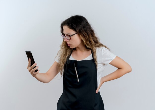 Donna giovane parrucchiere professionista in grembiule che tiene smartphone guardando lo schermo confuso e scontento in piedi sopra il muro bianco