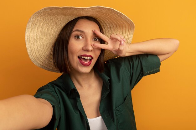 Donna graziosa allegra con il segno della mano di vittoria di gesti del cappello della spiaggia isolato sulla parete arancio