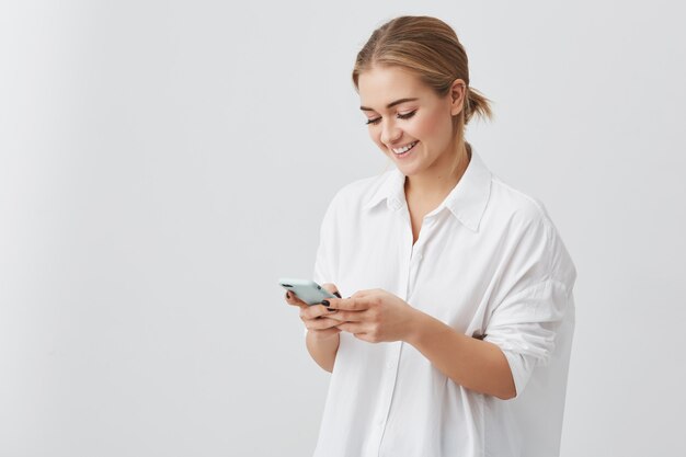 Donna graziosa attraente con capelli biondi in camicia bianca che sorride mentre per mezzo del telefono cellulare che chiacchiera con la sua posa del ragazzo. Concetto di bellezza e giovinezza