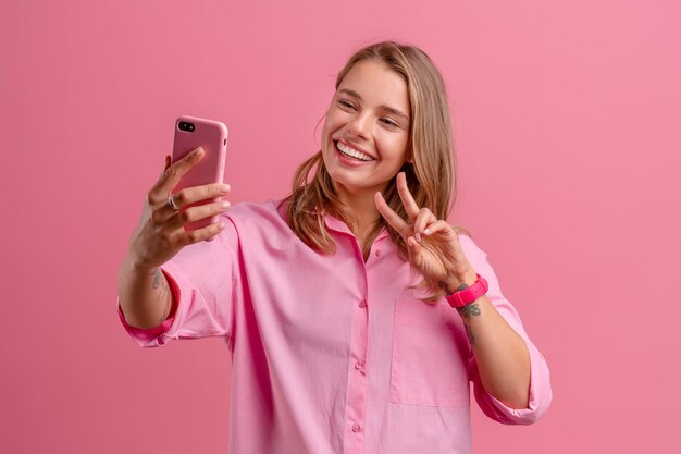 Donna graziosa bionda in camicia rosa che tiene sorridente che tiene facendo uso dello smartphone