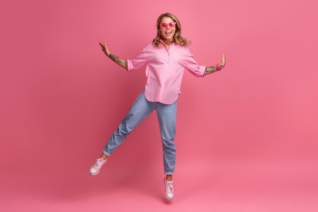 Donna graziosa bionda in camicia rosa e jeans sorridente che salta su sfondo rosa isolato divertendosi