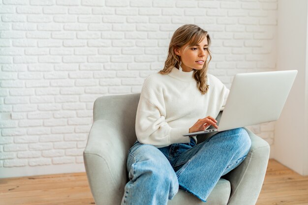 Donna graziosa con i capelli ondulati biondi che si siede sul sofà a casa che lavora al computer portatile