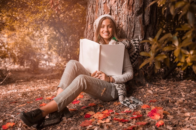 Donna graziosa con il libro che si siede vicino all&#39;albero