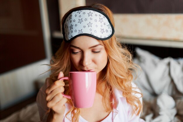 Donna graziosa con il sorriso schietto che posa nella maschera per gli occhi nella sua camera da letto e che beve tè caldo.