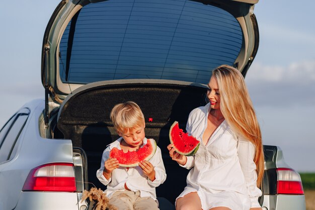 Donna graziosa dei capelli biondi con il piccolo figlio biondo al tramonto che si rilassa dietro l'automobile e che mangia anguria. estate, viaggi, natura e aria fresca in campagna.