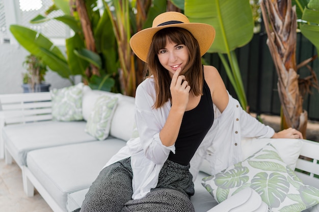 Donna graziosa del brunette in cappello di paglia che si siede sulla terrazza tropicale accogliente