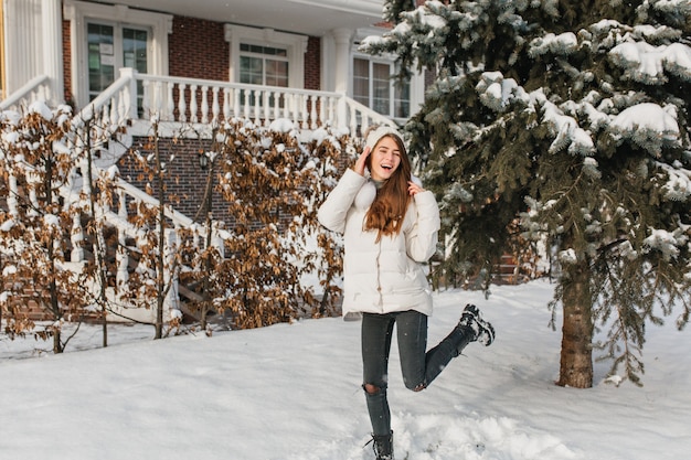 Donna graziosa in jeans strappati che ballano sulla strada innevata nella giornata invernale. Ritratto all'aperto di raffinata donna europea in giacca bianca che scherza nel cortile accanto all'abete rosso.