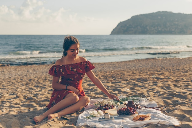 Donna graziosa in vestito rosso che si siede sulla spiaggia durante il giorno