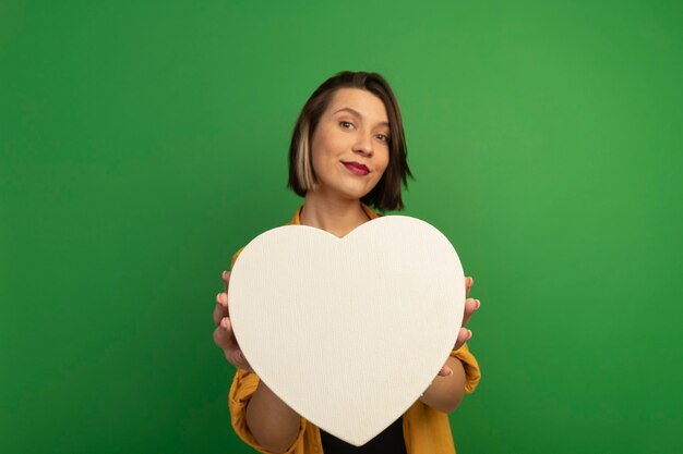 Donna graziosa soddisfatta che tiene forma del cuore isolata sulla parete verde