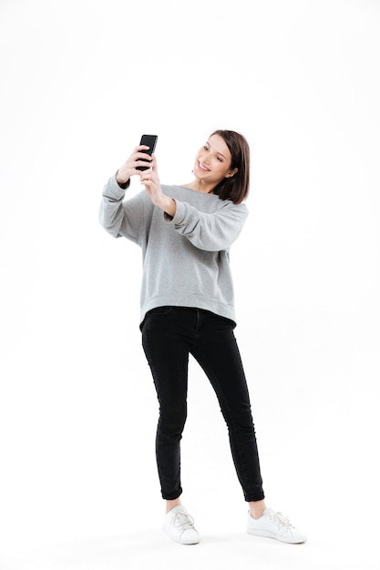 Donna graziosa sorridente che sta e che prende selfie sul telefono cellulare