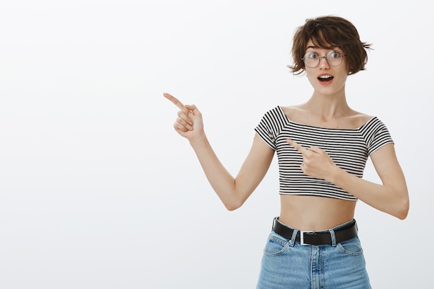 Donna hipster divertita con gli occhiali che punta le dita nell'angolo superiore sinistro al tuo logo o banner per scoprire i dettagli