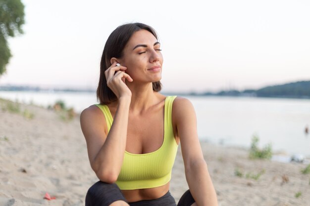 donna in abbigliamento sportivo al tramonto sulla spiaggia della città che riposa dopo l'allenamento ascoltando musica in cuffie wireless