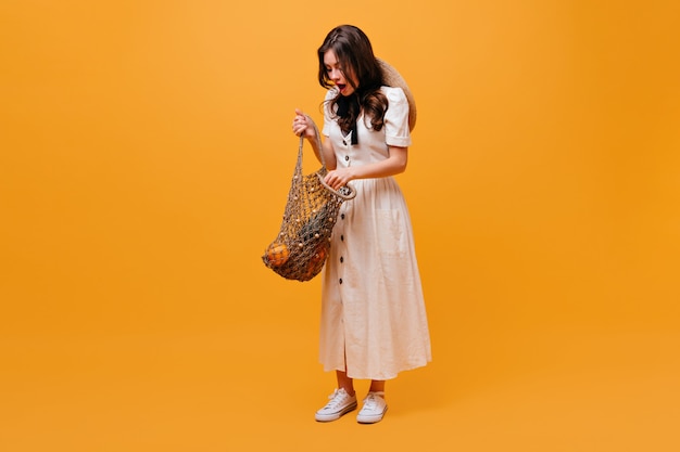 Donna in abito bianco apre la borsa della spesa con frutta e pose su sfondo arancione.