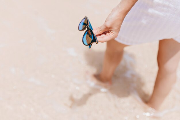 Donna in abito bianco in piedi in acqua al resort. Foto del modello femminile abbronzato che tiene gli occhiali da sole in mano.