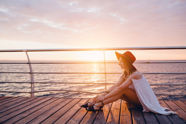 donna in abito bianco seduto in riva al mare all'alba in stato d'animo romantico indossando il cappello rosso