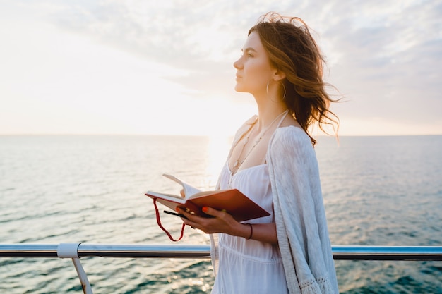 donna in abito estivo bianco che cammina in riva al mare all'alba con il libro del diario in stato d'animo romantico, pensando e prendendo appunti